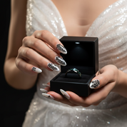 A close-up lifestyle shot of a person in a sparkling white dress holding an open black ring box. Inside the box is a ring with a dark green stone and a silver band.