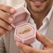A close-up of a man holding an open pink velvet ring box, showcasing a yellow gold eternity ring set with small clear stones. The man is wearing a white collared shirt and a light beige blazer.