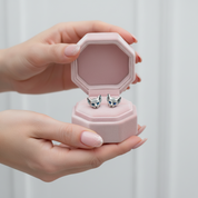 A pair of silver fox-head stud earrings with blue gemstone eyes displayed in an open pink octagonal jewelry box, held by two hands with glittery pink nails against a soft light gray background.