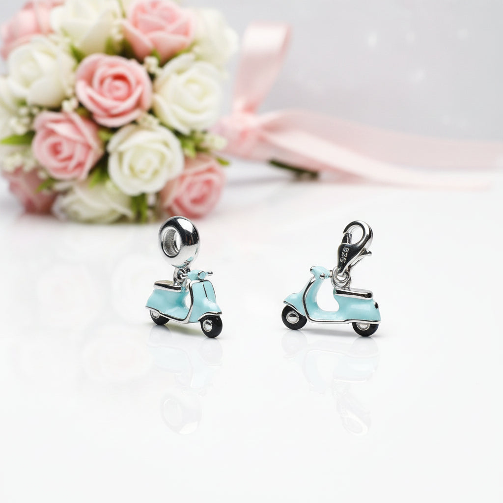 Two small light blue scooter charms with silver-tone metal hardware and black wheels, displayed on a white reflective surface with a blurred bouquet of pink and white roses in the background.