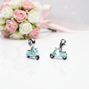 Two small light blue scooter charms with silver-tone metal hardware and black wheels, displayed on a white reflective surface with a blurred bouquet of pink and white roses in the background.