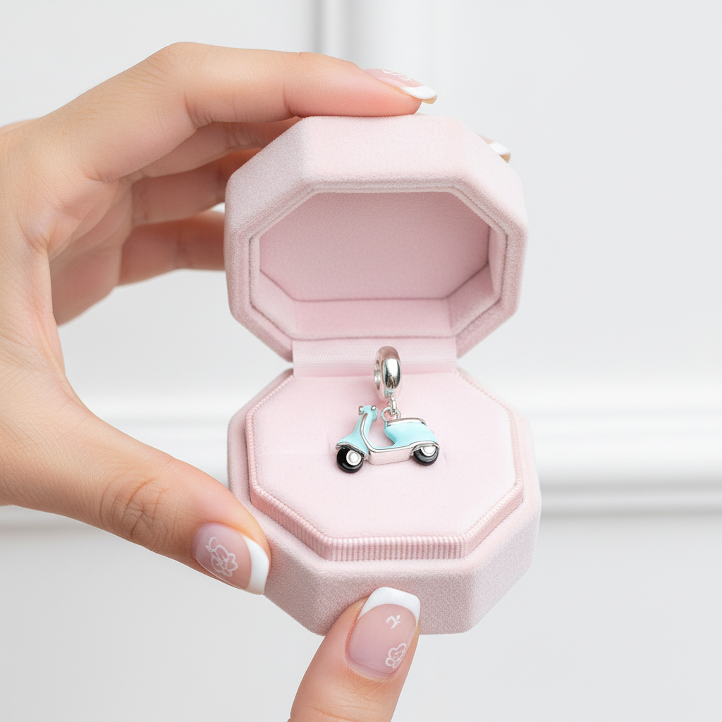 A hand with decorated nails holds an open pink jewelry box containing a small silver-tone charm shaped like a light blue scooter.