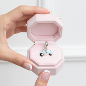 A hand with decorated nails holds an open pink jewelry box containing a small silver-tone charm shaped like a light blue scooter.