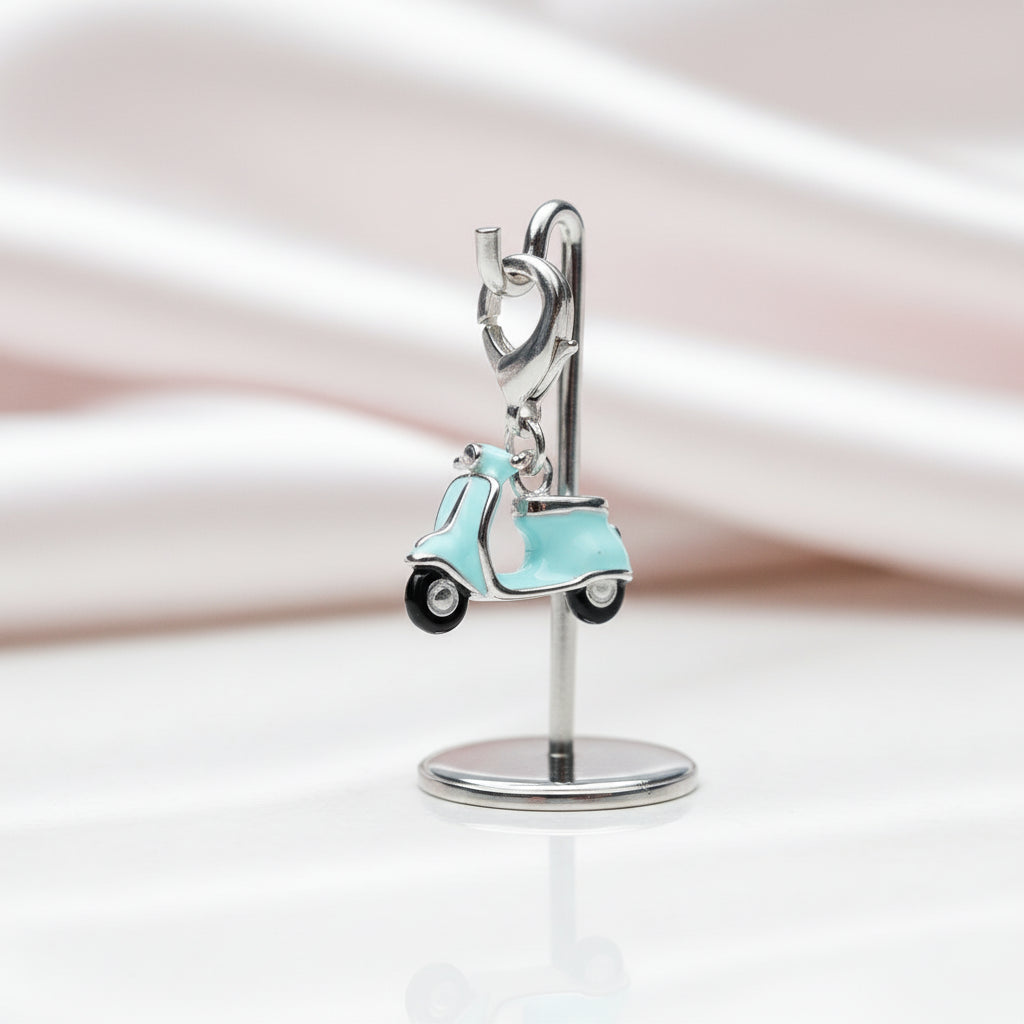 A silver-tone jewelry display stand holding a small charm shaped like a light aqua scooter with black wheels, photographed on a soft white and pale pink satin background.