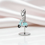 A silver-tone jewelry display stand holding a small charm shaped like a light aqua scooter with black wheels, photographed on a soft white and pale pink satin background.