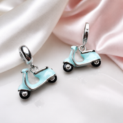 A pair of silver-tone scooter charms with light blue enamel, displayed on a white surface with draped pink and white satin fabric.