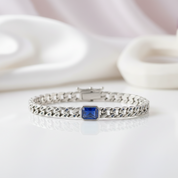 A silver-toned chain bracelet featuring a square-cut blue gemstone centerpiece, displayed on a clean white reflective surface with soft, out-of-focus white decorative objects in the background.