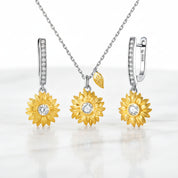 A jewelry set featuring a necklace and matching earrings with gold sunflower pendants and silver-toned hoops set with small clear stones, displayed on a white marble surface.