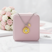 A gold and silver necklace with a sunflower pendant and a small leaf charm, displayed on a pink jewelry box with pink and white roses in the background.