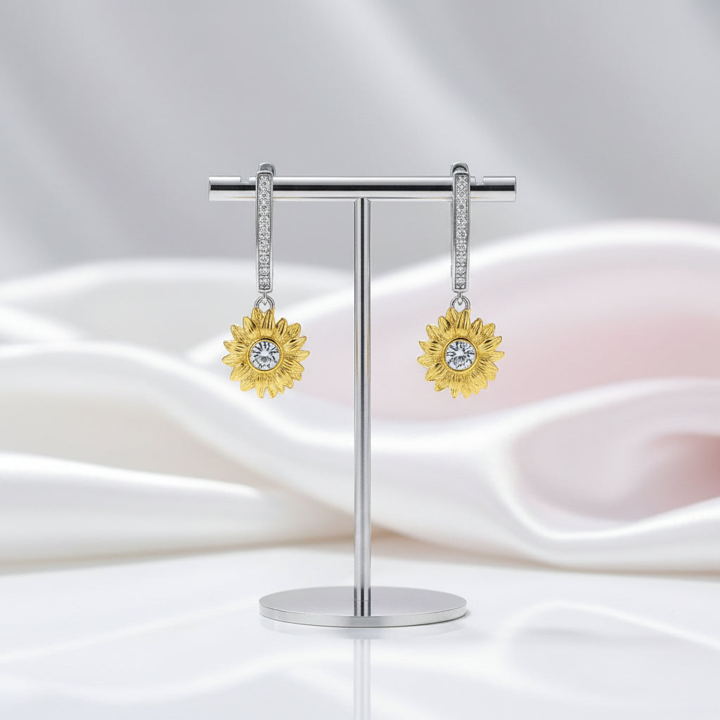 A pair of gold flower-shaped drop earrings with clear gemstones, displayed on a silver stand against a soft white and pale pink draped fabric background.