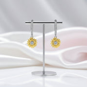 A pair of gold flower-shaped drop earrings with clear gemstones, displayed on a silver stand against a soft white and pale pink draped fabric background.