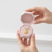 A person holds an open octagonal pink jewelry box containing a gold pendant necklace with a flower and heart charm, set against a clean white wall.
