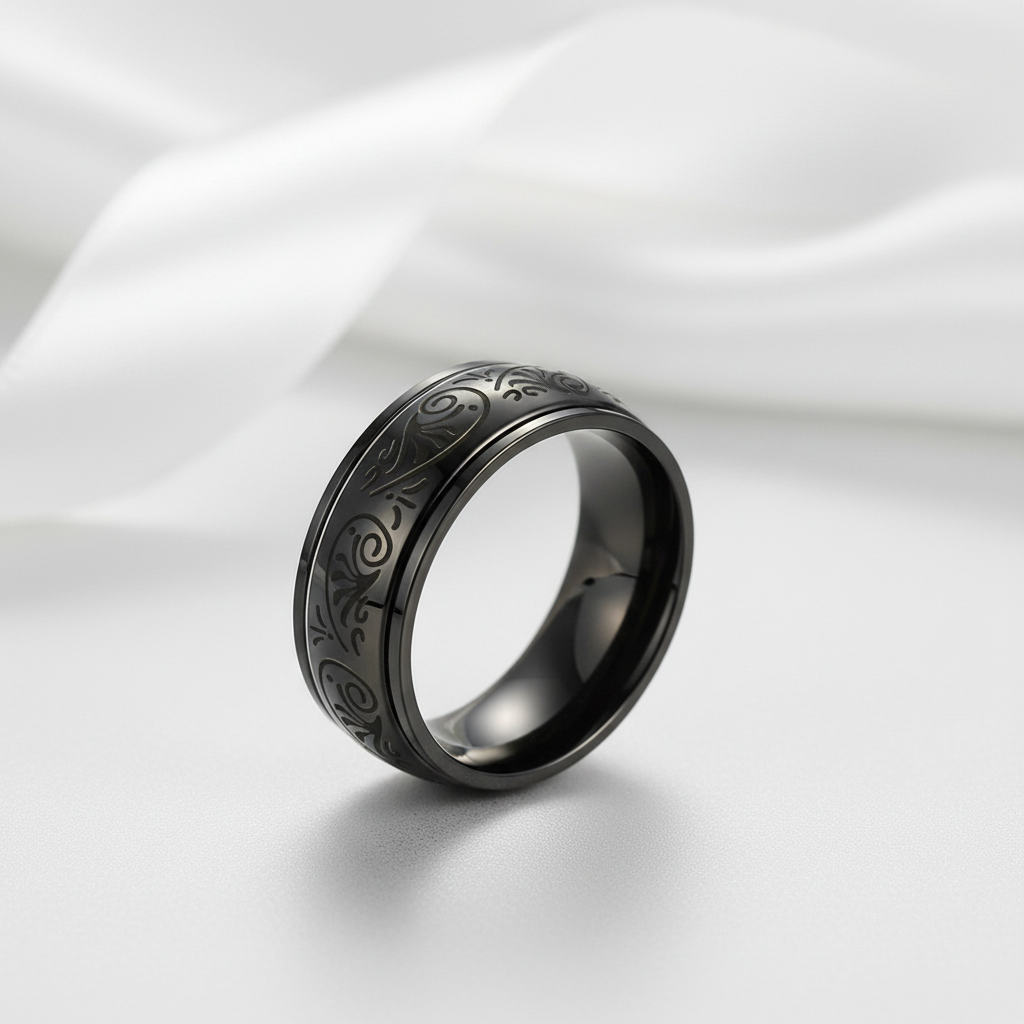 A black metallic band ring with engraved swirling patterns, displayed on a soft white satin fabric background.