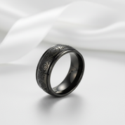 A black metallic band ring with engraved swirling patterns, displayed on a soft white satin fabric background.