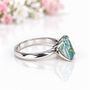 A silver ring featuring a marquise-cut iridescent green gemstone, displayed on a reflective white surface with soft pink and white flowers blurred in the background.