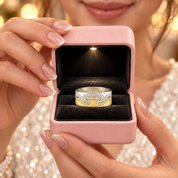 A close-up lifestyle shot of a woman holding an open pink ring box. Inside the box is a two-tone ring with a silver band, a gold wave-like inlay, and a row of clear stones.