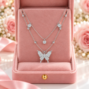 A double-strand silver necklace set with a butterfly pendant and small round gemstone accents, presented in an open pink velvet jewelry box with blurred pink roses in the background.