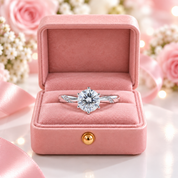 A silver solitaire engagement ring with a round clear gemstone is displayed inside an open pink velvet jewelry box, surrounded by soft pink roses, white baby's breath, and pink ribbon on a white surface with warm bokeh lights.