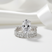 An oval-cut diamond engagement ring with a silver-toned band, shown with a matching diamond-studded wedding band on a clean white background.