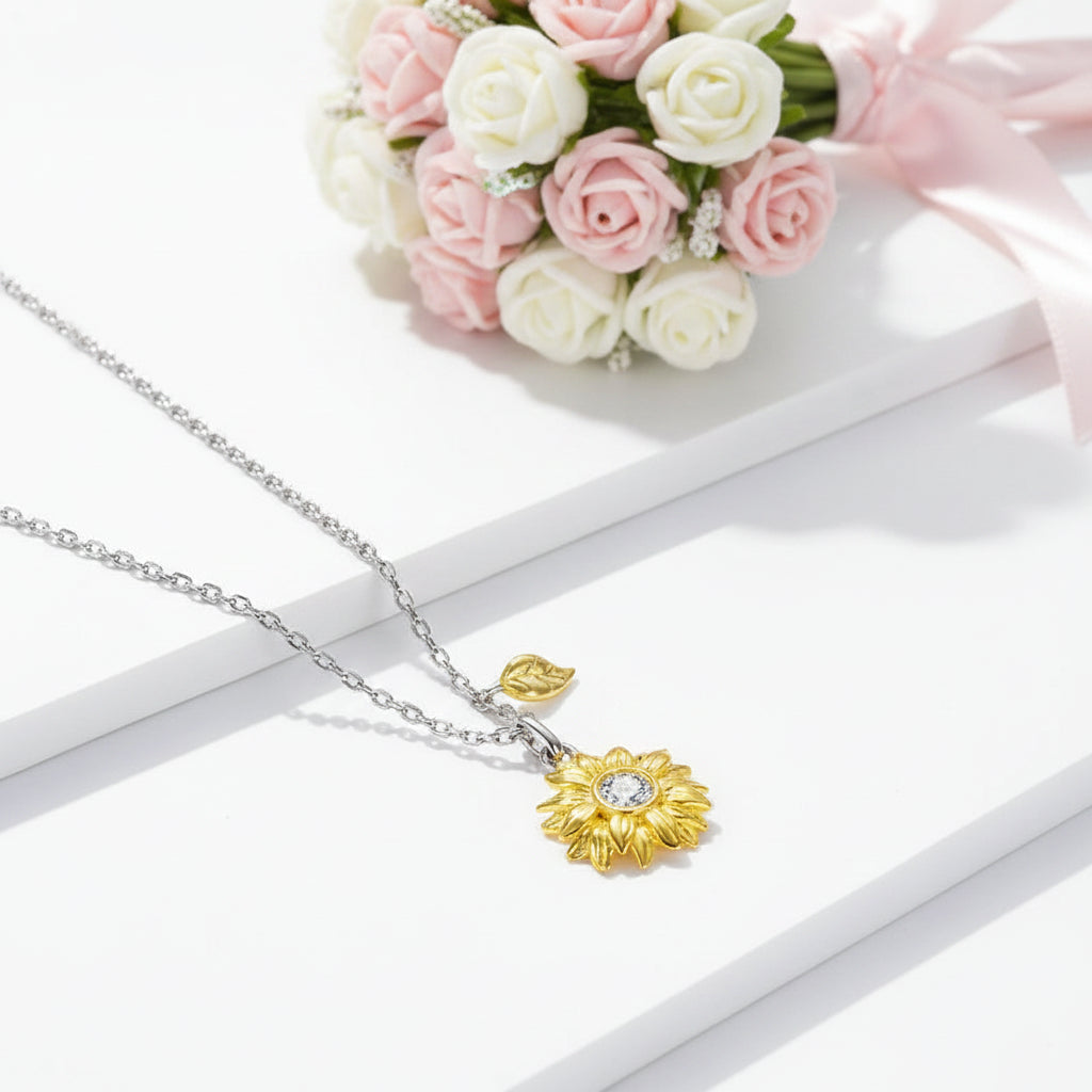 A delicate necklace with a yellow gold flower pendant and a small gold leaf charm on a silver chain, styled on white display blocks with pink and white roses in the background.
