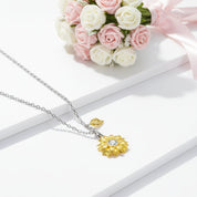 A delicate necklace with a yellow gold flower pendant and a small gold leaf charm on a silver chain, styled on white display blocks with pink and white roses in the background.