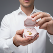 A man in a white dress shirt holds an open light pink jewelry box displaying a gold flower-shaped pendant necklace on a gray studio background.