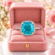 A cushion-cut turquoise-blue gemstone ring with a diamond halo and diamond-set band, displayed in an open pink velvet ring box with a pink ribbon, set against a soft-focus floral background.