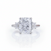 A radiant cut diamond ring with baguette side stones in a silver-toned setting, centered on a reflective white surface.