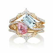 A luxury multi-band ring featuring a large light blue emerald-cut center stone, a pink pear-cut accent stone, and a small diamond, set in gold and white gold bands paved with clear diamonds, on a white background with a soft reflection.