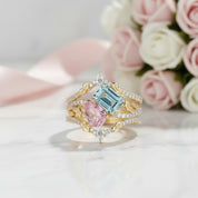 A gold ring with a large light blue emerald-cut center stone, a pink oval center stone, and small clear stones, displayed on a white marble surface with pink and white roses in the background.