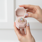 A close-up of a hand holding an open light pink jewelry box containing a gold ring with a large multicolor gemstone, set against a soft white background.
