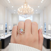 A close-up of a hand wearing a gold ring with a light blue rectangular gemstone and a pink marquise-cut gemstone, set against a blurred luxury jewelry store background with a chandelier.