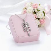 A silver dreamcatcher pendant necklace with clear crystals, displayed on a pink jewelry box with a bouquet of pink and white roses in the background.