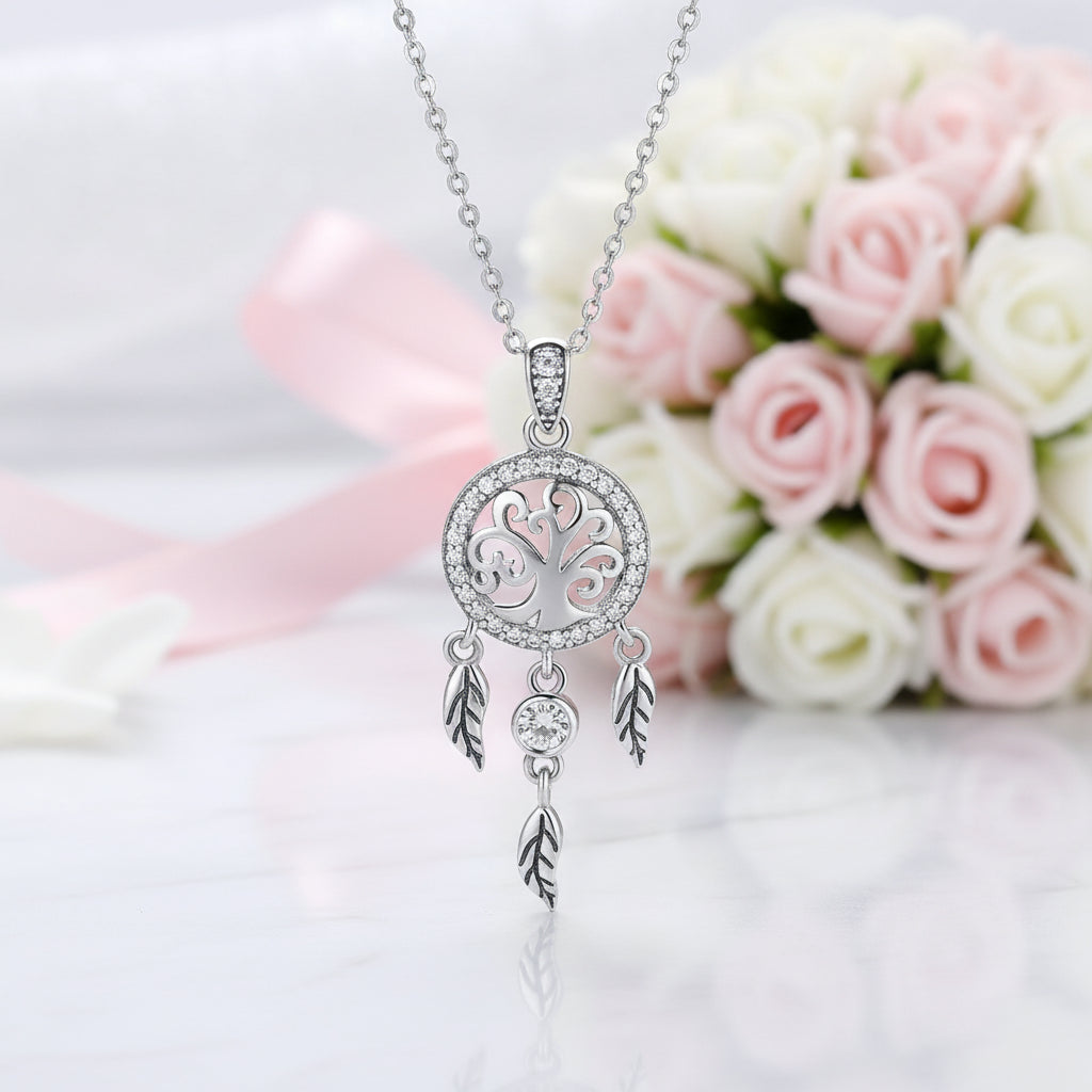 A silver dreamcatcher pendant necklace with a tree of life design and dangling feathers, displayed against a soft-focus background of pink and white roses.