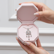 A silver dreamcatcher pendant necklace displayed inside an open octagonal pink jewelry box, held by two hands with manicured nails.