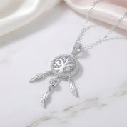 A silver dreamcatcher pendant necklace with a tree of life design and dangling feathers, displayed on a white marble surface with soft pink satin fabric in the background.