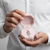 A silver-toned pendant necklace with a crescent moon and small clear stones is displayed inside an open light pink jewelry box, held by a person wearing a white dress shirt.