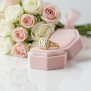 A gold ring with a rectangular clear gemstone and a small side diamond, displayed on a pink jewelry box with a bouquet of pink and white roses in the background.