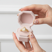 A gold ring with a teardrop-shaped clear gemstone is displayed inside an open light pink velvet ring box, held by two hands with floral nail art against a soft white paneled background.