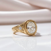 A yellow gold ring with a large oval clear gemstone and a smaller round diamond accent on the band, resting on a white reflective surface with soft pink fabric in the background.