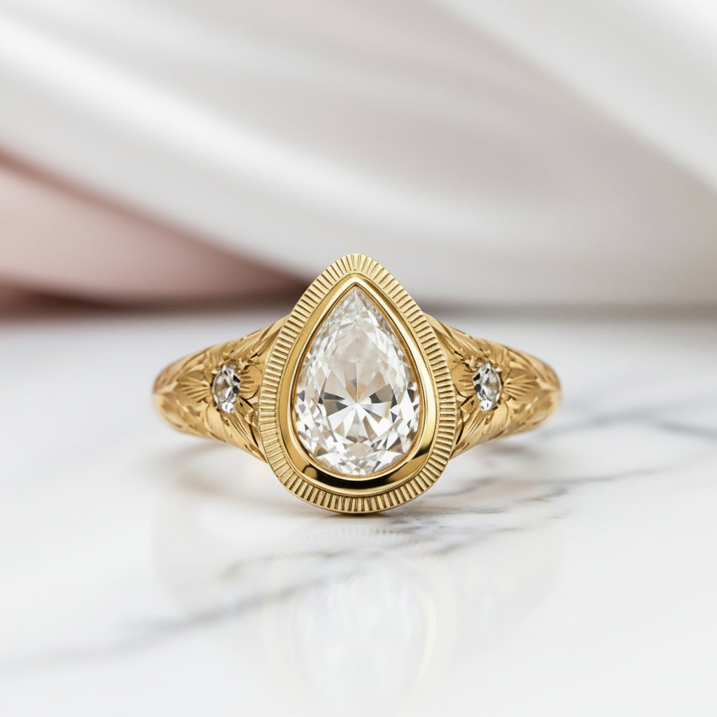 A gold ring with a pear-cut clear center stone and two small side stones, photographed on a light marble surface with soft draped fabric in the background.