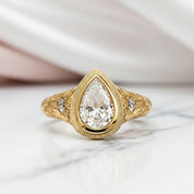 A gold ring with a pear-cut clear center stone and two small side stones, photographed on a light marble surface with soft draped fabric in the background.