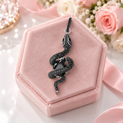 A black snake pendant with a central black gemstone, set in silver-toned metal and detailed with small black stones, shown on a clean white background with a soft reflection below.