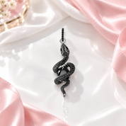 A black snake pendant with a central black gemstone, set in silver-toned metal and detailed with small black stones, shown on a clean white background with a soft reflection below.