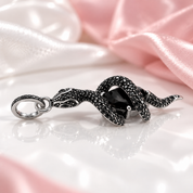 A black snake pendant with a central black gemstone, set in silver-toned metal and detailed with small black stones, shown on a clean white background with a soft reflection below.