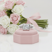 A radiant-cut diamond engagement ring with a silver-toned band, displayed on a pink velvet ring box with a bouquet of pink and white roses in the background.
