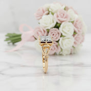 A yellow gold ring with a square-cut clear gemstone in a four-prong setting, displayed on a white marble surface with a blurred bouquet of pink and white roses in the background.