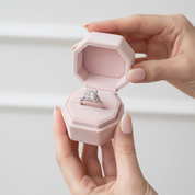 A silver ring with a large square-cut clear gemstone is displayed inside an open light pink velvet ring box, held by two hands against a clean white background.