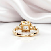 A gold ring with a geometric crown-like setting and a clear square gemstone, photographed on a white surface with soft draped satin fabric in the background.