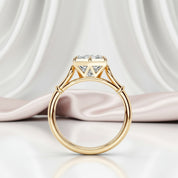 A yellow gold solitaire engagement ring with a large cushion-cut clear gemstone, displayed on a reflective white surface with draped light pink satin fabric in the background.